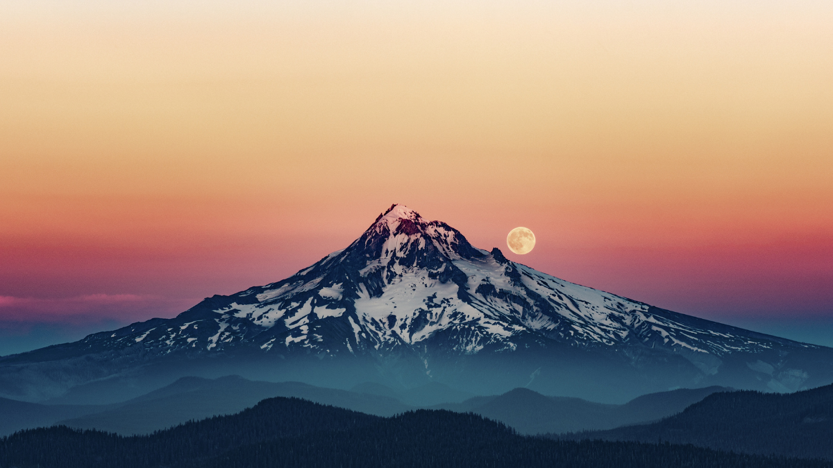 A spectacular photo of the moon rising over the righthand side of Mount Hood. The sky is a fiery gradient of  yellow fading down to rosy purple